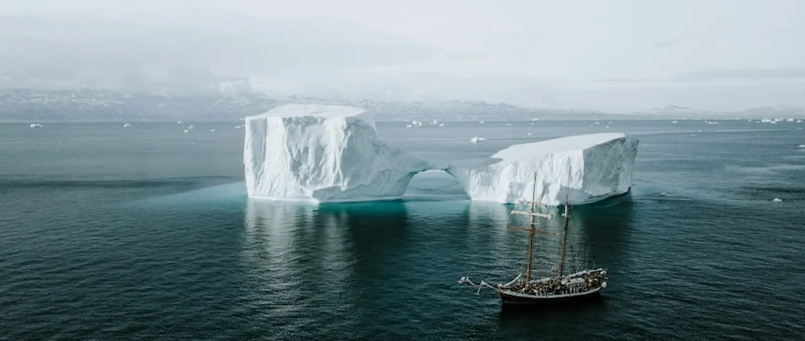 A small sailing vessel beside two icebergs in flat light.